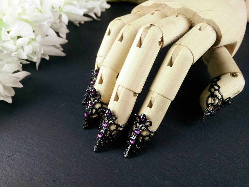 Puede incluir: Cuatro anillos de encaje negro para la punta de los dedos con diamantes de imitaci&oacute;n morados. Los anillos est&aacute;n en un maniqu&iacute; de mano de madera.