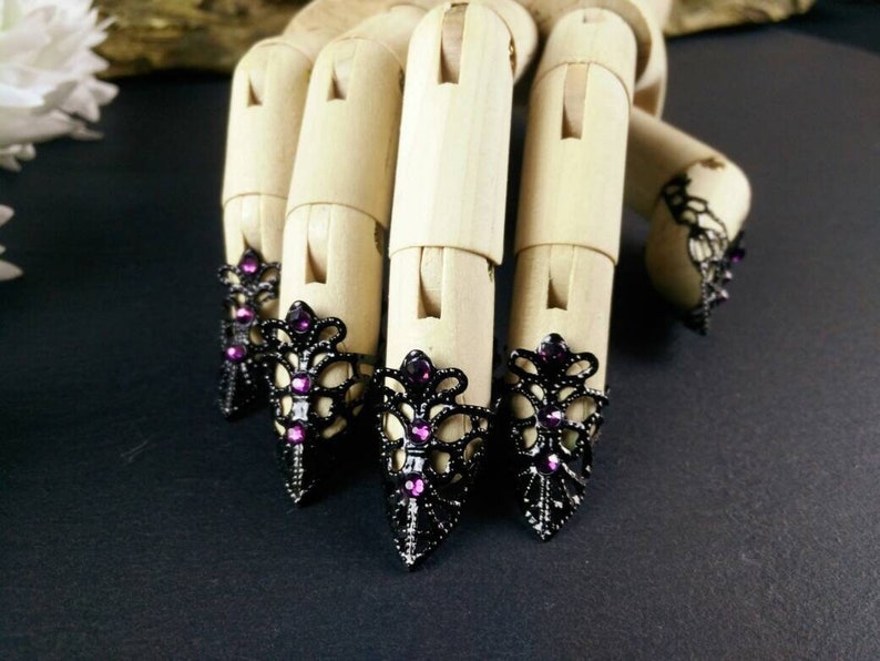 Puede incluir: Cinco anillos de dedo de metal negro con un delicado dise&ntilde;o de encaje y diamantes de imitaci&oacute;n morados. Los anillos se llevan en un maniqu&iacute; de mano de madera.