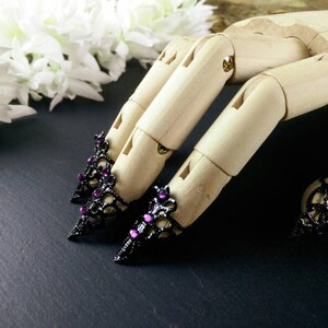 Puede incluir: Tres anillos de dedo negros y morados con joyas y un dise&ntilde;o de encaje. Los anillos est&aacute;n en un modelo de mano de madera.