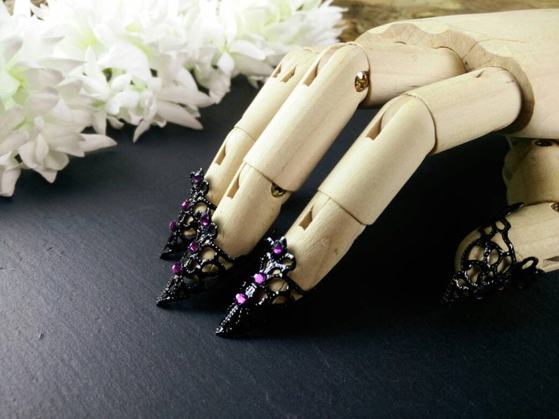 Puede incluir: Tres anillos de dedo negros y morados con joyas y un dise&ntilde;o de encaje. Los anillos est&aacute;n en un modelo de mano de madera.