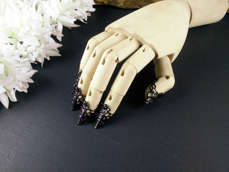 Puede incluir: Un maniqu&iacute; de mano de madera que lleva anillos de encaje negro para la punta de los dedos con diamantes de imitaci&oacute;n morados. Los anillos son puntiagudos y tienen un estilo g&oacute;tico.