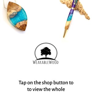 Op de afbeelding: Een teal- en paarse harspen met een gouden rand en een houten basis. De pen rust op een stuk hout. De afbeelding toont ook een teal- en paarse harshanger met een houten basis. De tekst "WEARABLEWOOD" is zichtbaar onder de afbeelding.