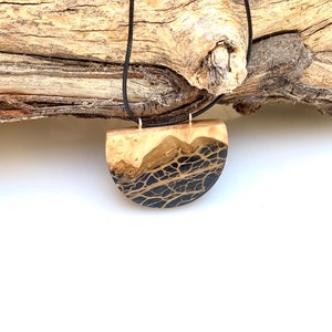 May include: A black and gold resin pendant necklace with a half-circle shape. The pendant is made of wood and resin and has a natural wood grain pattern. The necklace is on a black cord.