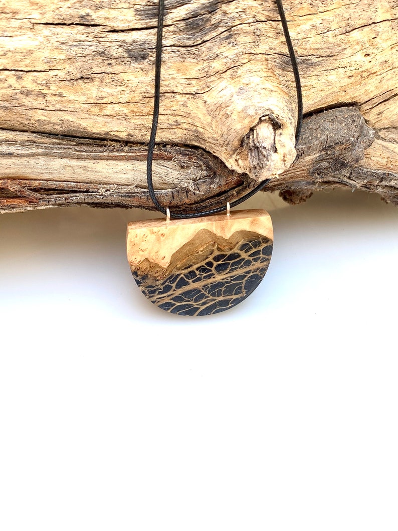 May include: A black and gold resin pendant necklace with a half-circle shape. The pendant is made of wood and resin and has a natural wood grain pattern. The necklace is on a black cord.
