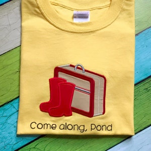 May include: Yellow t-shirt with embroidered red rain boots and a brown suitcase. The text "Come along, Pond" is embroidered below the image.