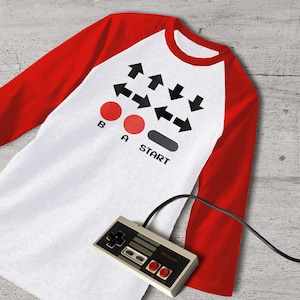 May include: A white and red baseball t-shirt with a black and white video game controller graphic on the front. The graphic includes arrows, a start button, and two red buttons labelled "B" and "A".