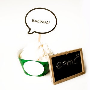 May include: A green and white cupcake liner with white frosting and a speech bubble that says "Bazinga!" on a stick. A small black chalkboard with the equation "e=mc²" written in white chalk is next to the cupcake.