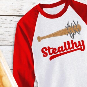 May include: A white and red baseball-style shirt featuring a wooden baseball bat graphic with nails. The word "Stealthy" is written in red script below the bat. A wooden baseball bat is in the background.