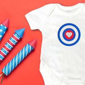 May include: Four red, white, and blue paper firecrackers with a white baby onesie with a red, white, and blue target with a white heart in the center on a red background.