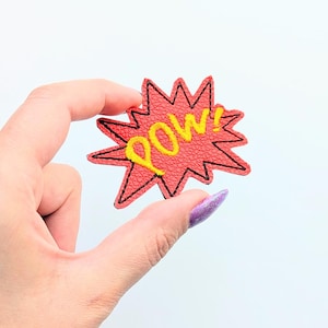 Può includere: Una toppa rossa a forma di stella con la scritta "POW" in giallo, bordata di nero. La toppa è tenuta su uno sfondo bianco. La toppa è un design in stile fumetto.