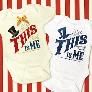 May include: Two white baby onesies with short sleeves. One onesie has a red "This is Me" graphic with a black top hat and a gold star. The other onesie has a blue "This is Me" graphic with a black top hat and a gold star.