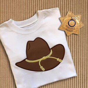 May include: White t-shirt with a brown cowboy hat embroidered on the front. The hat has a yellow band and a brown brim. The shirt also has a gold sheriff's star badge with the text "King County Sheriff" embroidered on the back.