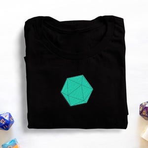 May include: Black t-shirt featuring a teal geometric design, with various coloured dice scattered around. The design is a teal, multi-faceted shape. The dice are different colours and shapes.
