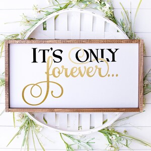 May include: A white wooden sign with a black and gold inscription that reads "It's only forever..." The sign is framed in a brown wood frame and is sitting on a white wooden surface.