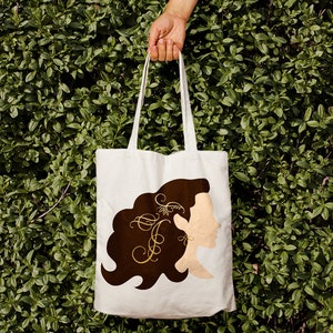 May include: A white tote bag with a brown silhouette of a woman's head and shoulders with a gold floral design in her hair. The design includes the letter "F" in a decorative font.