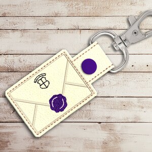 May include: A white leather keychain shaped like an envelope with a purple seal and a black and white crest. The keychain has a silver metal clasp.