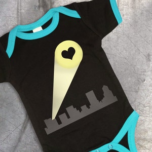Puede incluir: Un body negro para bebé con ribete turquesa. El body tiene un gráfico de un horizonte de ciudad con un foco amarillo que brilla sobre él. El foco tiene una forma de corazón en el centro.