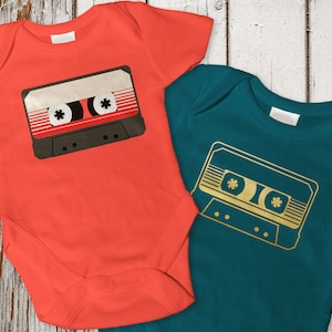 May include: Two baby onesies with a cassette tape design. The onesie on the left is orange with a black and red cassette tape. The onesie on the right is teal with a gold cassette tape.