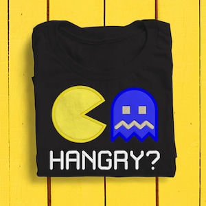 Puede incluir: Camiseta negra con un Pac-Man amarillo y un fantasma azul. El texto "HANGRY?" está impreso debajo del gráfico.