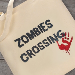 May include: A white canvas tote bag with the text "Zombies Crossing" in black embroidery. A red handprint with a few drops of blood is embroidered on the right side of the bag.