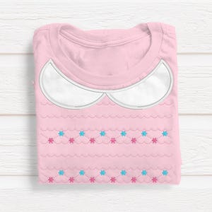 Könnte beinhalten: Ein gefaltetes rosa T-Shirt mit weißem Kragendesign. Das Shirt hat ein gewelltes Muster mit abwechselnd blauen und rosa floralen Akzenten. Das Shirt wird auf einer weißen Holzoberfläche präsentiert.