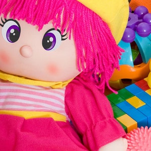 May include: A pink and yellow plush doll with long pink yarn hair and big purple eyes. The doll is surrounded by colorful toys, including a yellow rubber duck, a blue and orange toy, and a stack of colorful building blocks.