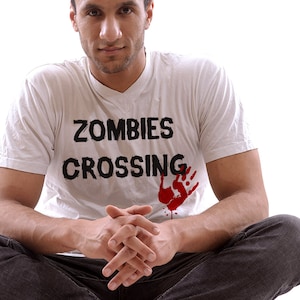 May include: A man wearing a white t-shirt with the text "Zombies Crossing" and a red handprint. He is sitting with his legs crossed and his hands clasped in his lap.