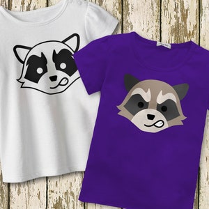 May include: Two t-shirts with a raccoon face graphic. The shirt on the left is white with a black raccoon face. The shirt on the right is purple with a brown raccoon face.