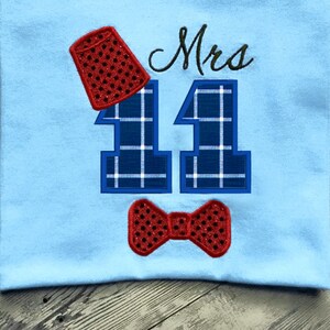 May include: A blue and white plaid number "11" with a red bow tie and a red hat with black polka dots. The text "Mrs" is embroidered above the number.