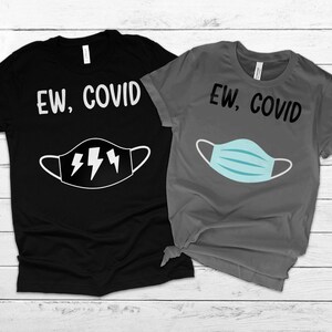 May include: Two t-shirts with the text "EW, COVID" printed on them. One shirt is black with a white lightning bolt mask, and the other shirt is grey with a blue medical mask.