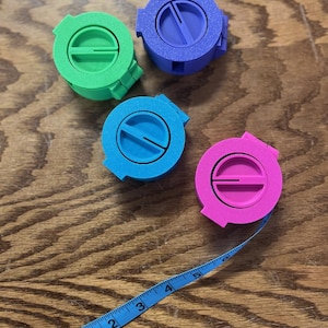 May include: Four colorful measuring tape holders in green, purple, blue, and pink. A blue measuring tape extends from the pink holder, showing measurements in inches. The holders are on a wooden surface.