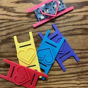 May include: A collection of colorful thread spool holders in red, yellow, blue, and purple, each with a heart-shaped design. One pink holder contains a small spool of fabric with a floral pattern. The items are arranged on a wooden surface.