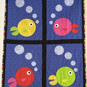 May include: A colorful, handmade quilt featuring four square panels with a blue background dotted with white. Each panel showcases a different colored fish: yellow, pink, red, and green, with white bubbles. The quilt has a black border and a multi-colored edge.