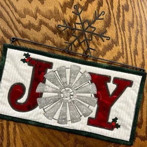 May include: A white fabric banner with the word "JOY" in red letters. The "O" is replaced with a silver windmill design. The banner is decorated with green holly leaves and red berries. A black metal snowflake is hanging above the banner.