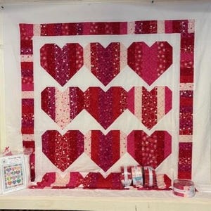 May include: A handmade quilt featuring nine large, heart-shaped designs in shades of red and pink. The hearts are pieced together with various patterned fabrics, set against a white background. Additional fabric rolls and a pattern are visible.