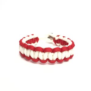 May include: A red and white paracord bracelet with a silver clasp. The bracelet is made of woven nylon cord in a repeating pattern of red and white. The bracelet is approximately 7 inches in length.