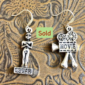 May include: A pair of silver earrings featuring a movie camera charm with the word "MOVIE" on it and an Oscar statue charm with the word "OSCAR" on it.