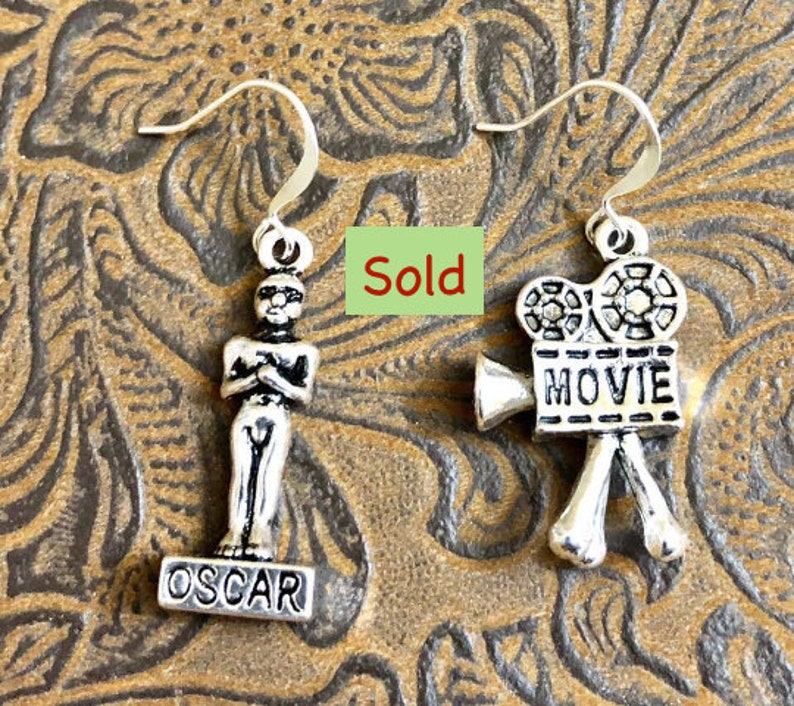 May include: A pair of silver earrings featuring a movie camera charm with the word "MOVIE" on it and an Oscar statue charm with the word "OSCAR" on it.