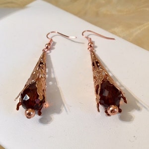 May include: A pair of copper-colored filigree earrings. Each earring features a cone-shaped design with a faceted, dark red crystal bead at the base. The earrings have a delicate, openwork pattern and are suspended from simple ear wires.