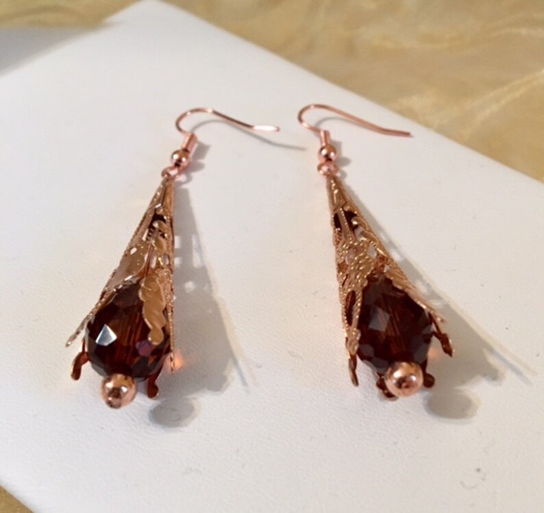 May include: A pair of copper-colored filigree earrings. Each earring features a cone-shaped design with a faceted, dark red crystal bead at the base. The earrings have a delicate, openwork pattern and are suspended from simple ear wires.