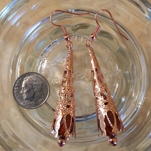 May include: A pair of copper-colored filigree earrings with a teardrop shape. The earrings have a detailed openwork design and are suspended from ear wires. A U.S. dime is next to the earrings for size comparison.