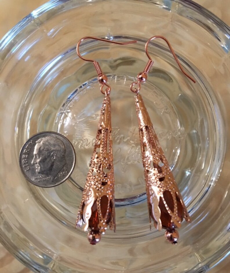 May include: A pair of copper-colored filigree earrings with a teardrop shape. The earrings have a detailed openwork design and are suspended from ear wires. A U.S. dime is next to the earrings for size comparison.