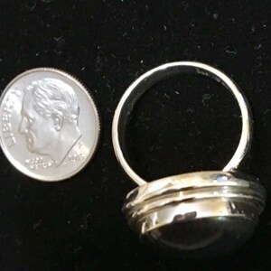 Op de afbeelding: Een zilveren ring met een ronde, donkergrijze steen in een bezel-setting. De ring heeft een brede band met een licht gewelfde bovenkant.