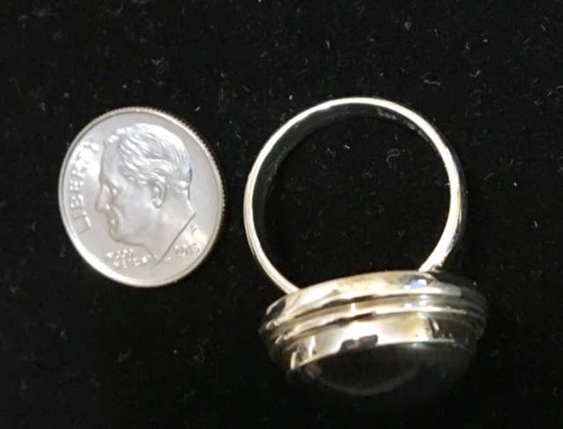Op de afbeelding: Een zilveren ring met een ronde, donkergrijze steen in een bezel-setting. De ring heeft een brede band met een licht gewelfde bovenkant.