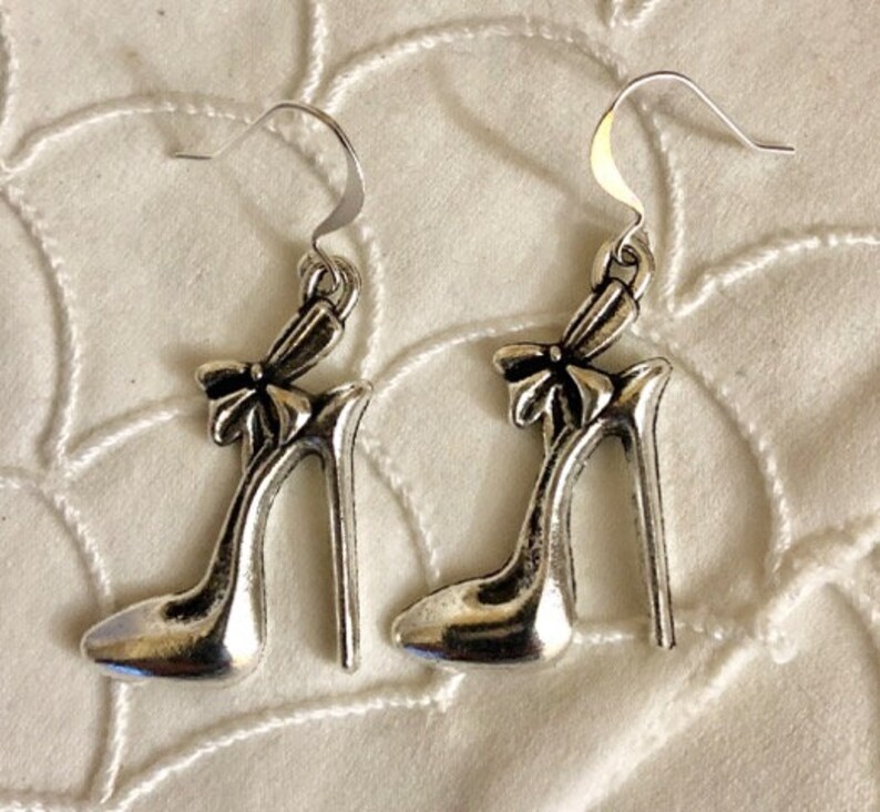 K&ouml;nnte beinhalten: Ein Paar silberne Ohrh&auml;nger mit einem Design einer High-Heel-Schuh mit einer Schleife oben.
