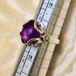 May include: A silver ring with a large, round, deep purple gemstone. The gemstone is set in a decorative silver setting with curved accents. The ring is being measured on a ring sizer.