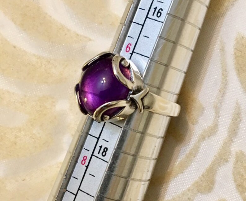 May include: A silver ring with a large, round, deep purple gemstone. The gemstone is set in a decorative silver setting with curved accents. The ring is being measured on a ring sizer.