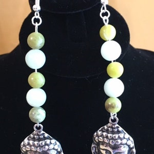 May include: A pair of dangle earrings featuring a silver Buddha head pendant. The earrings have a series of green and white beads. The earrings are suspended from silver-colored ear wires.