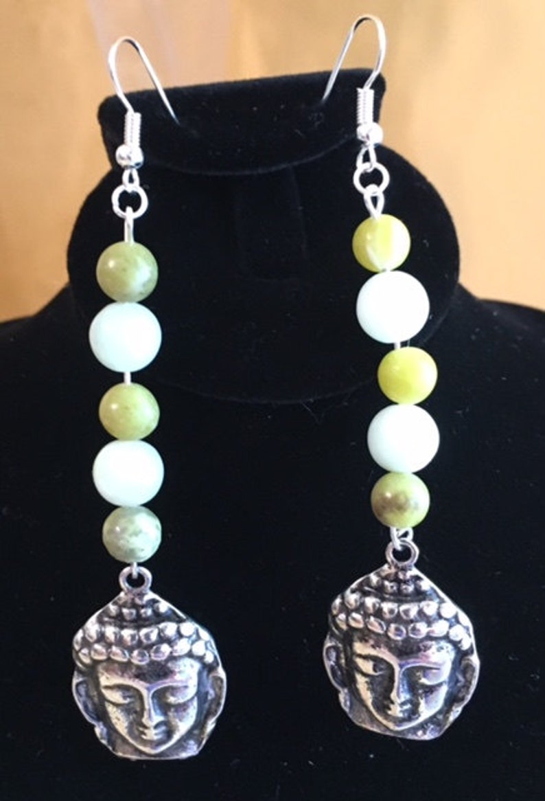 May include: A pair of dangle earrings featuring a silver Buddha head pendant. The earrings have a series of green and white beads. The earrings are suspended from silver-colored ear wires.