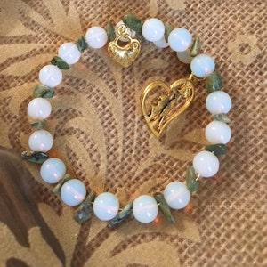 May include: A white and green beaded bracelet with two gold heart charms. The bracelet is made with white opal beads and green jade beads.